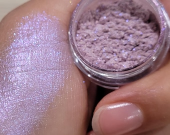 May include: A small jar of loose purple eyeshadow with a shimmery finish. The eyeshadow is shown on a person's hand.