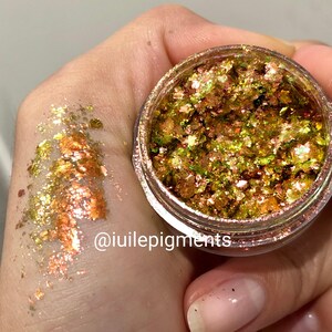 Könnte beinhalten: Ein kleines Glas mit schimmernden grünen und goldenen Glitterflocken. Der Glitter ist lose und hat einen metallischen Glanz. Das Glas ist mit dem Text "@iuiilepigments" beschriftet.