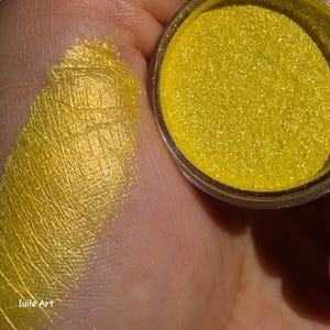 May include: A small round container filled with vibrant yellow metallic pigment powder, accompanied by a swatch of the same shimmering yellow powder applied to skin. The text "Iuile Art" is visible in the lower left corner of the image.