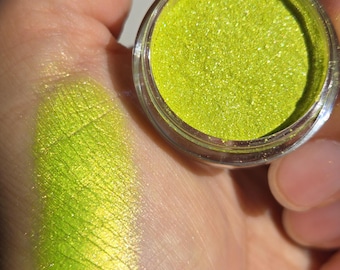 Könnte beinhalten: Ein kleines, durchsichtiges, rundes Döschen gefüllt mit leuchtend limettengrünem, schimmerndem Pigmentpuder. Ein Aufstrich desselben hellen, metallisch gelb-grünen Puders ist auf der Haut zu sehen und zeigt eine reflektierende Textur. Text: @iuilewatercolos.