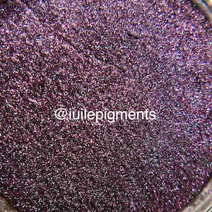 May include: Close-up of a shimmering, deep purple eyeshadow with a textured, granular appearance. The product has a metallic sheen and is densely packed. The text "@iuilepigments" is visible in white.