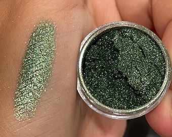 May include: A pot of dark green, shimmering eyeshadow next to a swatch of the same colour on a hand. The eyeshadow has a glittery, metallic finish. The swatch shows the texture and colour of the eyeshadow.