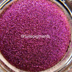 Puede incluir: Primer plano de un tarro lleno de purpurina magenta brillante. La purpurina tiene una textura fina y refleja la luz, creando un efecto brillante. El texto "@tuilepigments" es visible.