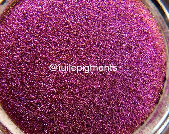 May include: Close-up of a jar filled with vibrant, sparkling magenta glitter. The glitter has a fine texture and reflects light, creating a shimmering effect. The text "@tuilepigments" is visible on the surface.