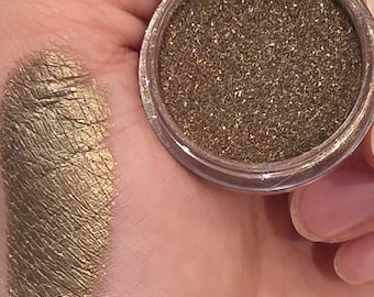 Könnte beinhalten: Nahaufnahme einer Hand, die ein kleines, klares Glas mit schimmerndem, goldbraunem Lidschatten hält. Links ist ein Farbmuster des gleichen Lidschattens mit Metallic-Finish zu sehen. Der Text "Iuile Art" ist auf dem Farbmuster zu sehen.