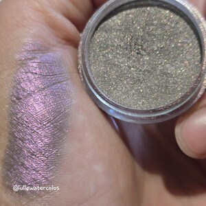 May include: A round container of loose, shimmering taupe-toned eyeshadow with a metallic finish. Next to the container is a swatch of the product, which displays a vibrant, iridescent purple shift with a fine, reflective glitter texture.
