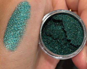 May include: A small jar of green glitter with a scoop of the glitter on a hand. The glitter is a dark green with a slight shimmer.