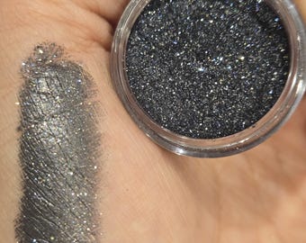 May include: A small clear circular container filled with dark charcoal gray glitter pigment, accompanied by a swatch of the same shimmering metallic gray powder applied to the skin, featuring the text iuilewatercolos.