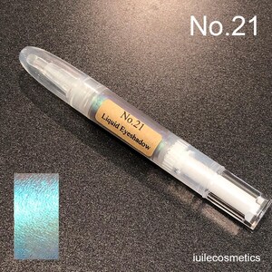 Könnte beinhalten: Ein transparentes Plastikröhrchen mit einem weißen Applikator-Spitz. Das Röhrchen ist mit "No. 21 Liquid Eyeshadow" beschriftet. Ein Abstrich des Lidschattens ist in der linken unteren Ecke zu sehen, der hellblau mit einem grün schimmernden Schimmer ist.