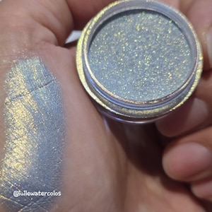 May include: A small round container filled with iridescent light blue and gold shimmer powder, accompanied by a swatch of the same pigment on skin. The text "@iuilewatercolos" is visible in the lower left corner of the image.