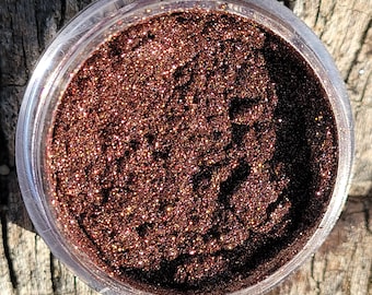 May include: A clear plastic jar filled with sparkling, dark brown glitter. The glitter has a textured appearance and reflects light, creating a shimmering effect. The jar is sitting on a weathered wooden surface.
