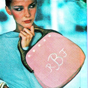 May include: A pink handbag with a wooden handle and white embroidered initials "RBF". The bag has a square shape with rounded corners and white button details. The bag is held by a person.
