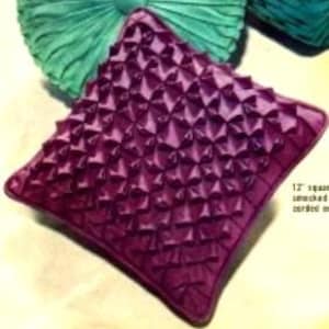 May include: A square, plum-colored decorative pillow with a textured, geometric design. The pillow is approximately 12 inches square. Two other teal-colored pillows are visible in the background.