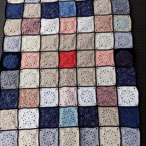 May include: A handmade crochet blanket featuring a patchwork design. The blanket is composed of numerous square blocks in various colors, including white, blue, pink, and beige, with a striking red square at the center. The squares are bordered with black yarn.