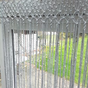 May include: A gray macrame curtain hangs from a wooden rod. The curtain features a woven top section with a diamond pattern, transitioning into vertical strands. The background shows a view of a green lawn and a white building.