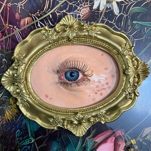 May include: A close-up of a painted eye with eyelashes, framed in a gold oval frame. The eye is surrounded by a light pink background with small pink dots.
