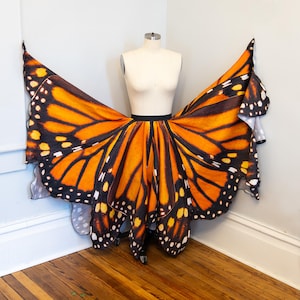 Puede incluir: Una falda con forma de alas de mariposa monarca, exhibida en un maniquí. La falda presenta un vibrante patrón naranja y negro, que imita las alas de una mariposa monarca. La falda está diseñada para envolverse alrededor de la cintura, con las puntas de las alas extendiéndose hacia afuera.
