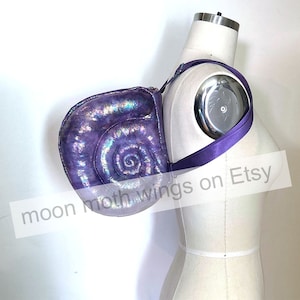 May include: A purple iridescent shell-shaped bag with a purple strap. The bag is worn on a mannequin's shoulder. The text "moon moth wings on Etsy" is visible in the image.