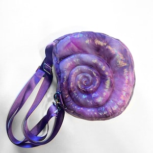 May include: A purple seashell-shaped crossbody bag. The bag has a spiral design and an iridescent finish. It features a matching purple adjustable strap and metal hardware.