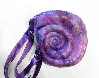 May include: A purple crossbody bag shaped like a seashell. The bag has a spiral design and a shimmering, iridescent finish. It features a matching purple adjustable strap and metal hardware.