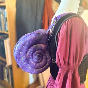 May include: A purple iridescent shell-shaped bag with a black strap. The bag is worn on a mannequin with a red dress.