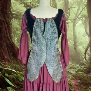 Puede incluir: Un vestido burdeos con mangas largas y falda escalonada, combinado con un corpiño negro y grandes alas de libélula iridiscentes. Las alas están detalladas con una red de venas y tienen un degradado de colores.