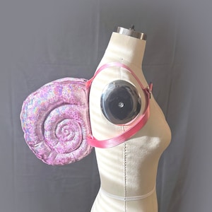 May include: A pink iridescent seashell-shaped backpack with a pink adjustable strap. The backpack is worn on a white dress form.