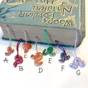 May include: Seven colorful snake bookmarks with different designs. The bookmarks are hanging from a book with the title "Wood's Popular Natural History".