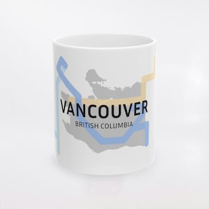 Op de afbeelding: Witte keramische mok met een kaart van Vancouver, British Columbia, en blauwe en gele lijnen. Het woord "VANCOUVER" is zwart gedrukt, met "BRITISH COLUMBIA" eronder. De mok is cilindrisch.