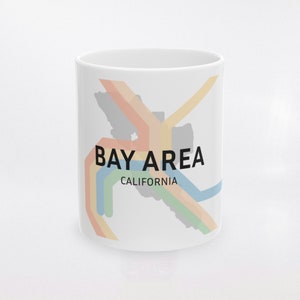 May include: White ceramic mug featuring the words "BAY AREA CALIFORNIA" in black, with a colorful map design in orange, yellow, green, and blue. The mug is a standard size and shape, suitable for hot or cold beverages.
