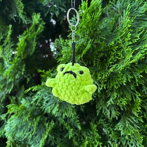 May include: A lime green crocheted frog keychain with a black stitched mouth and eyes. The frog has small arms and is suspended by a silver keyring and black cord. The background is a blurred view of green foliage.
