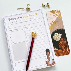 May include: A desk notepad with the text "Today is a new day" and a matching pen with a heart-shaped gold top. A bookmark with a woman's portrait and gold leaf accents is also present. Additional office supplies are visible.