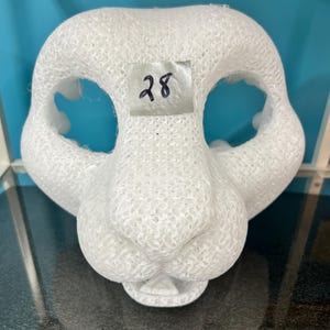 May include: A white 3D printed animal mask base with a mesh-like texture, featuring large eye openings, a prominent snout, and a small mouth area. A square piece of silver tape with the handwritten number 28 is attached to the forehead area.