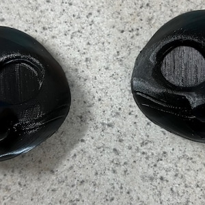 May include: Two black 3D-printed cat masks. Each mask features large eye openings, a nose, and a mouth. The masks are designed to fit the face and are likely for costume or cosplay purposes.