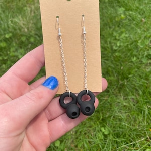 May include: A pair of black dangle earrings with a silver chain and a black circle charm. The earrings are hanging from a brown card.