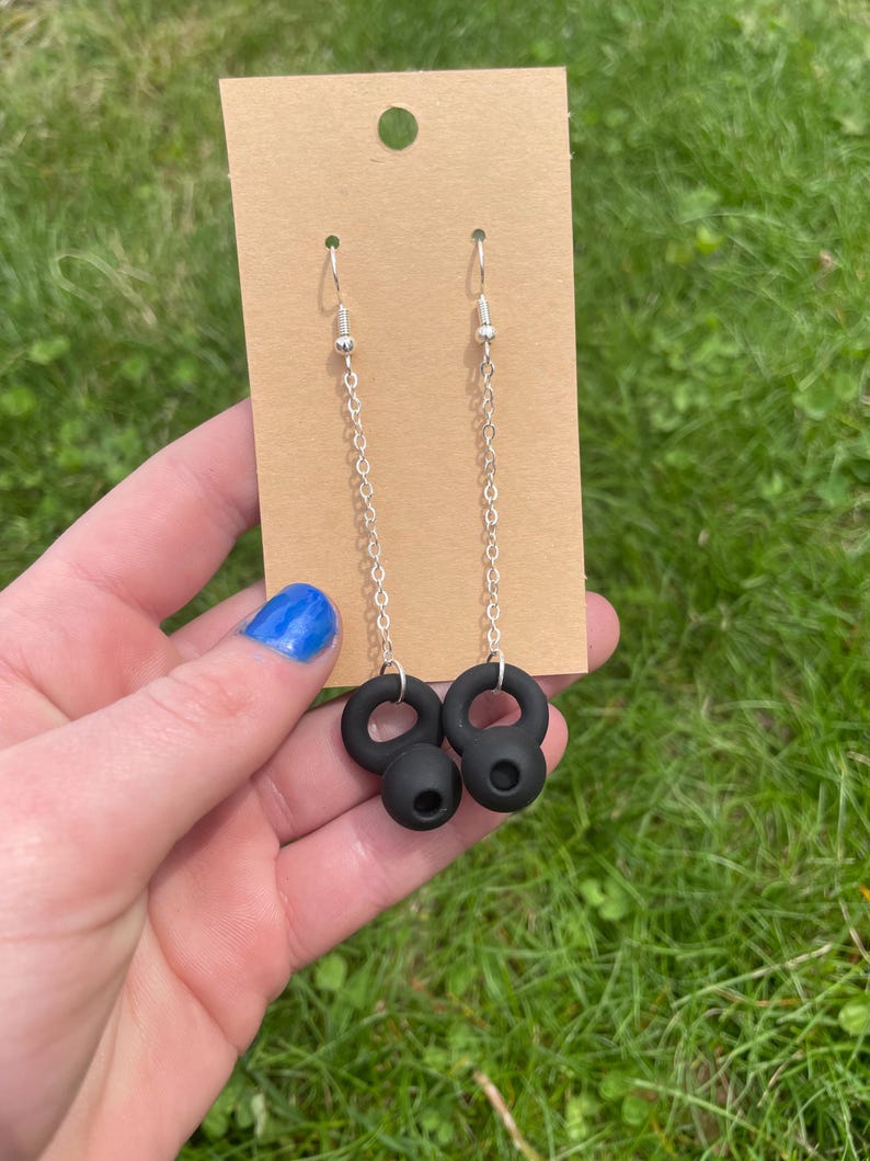 May include: A pair of dangle earrings featuring silver-toned metal chains attached to matte black kettlebell-shaped charms. The earrings are displayed on a rectangular tan cardboard backing card with a circular hole at the top for hanging.