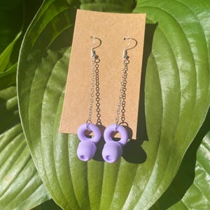 May include: A pair of dangle earrings featuring silver-toned metal chains attached to lavender-colored, ring-shaped charms. The earrings are displayed on a rectangular brown cardboard backing card with a circular hole at the top for hanging.