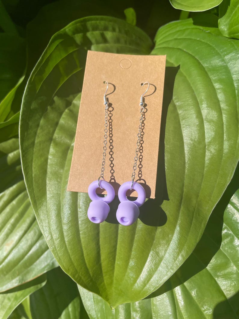 May include: A pair of dangle earrings featuring silver-toned metal chains attached to lavender-colored, ring-shaped charms. The earrings are displayed on a rectangular brown cardboard backing card with a circular hole at the top for hanging.
