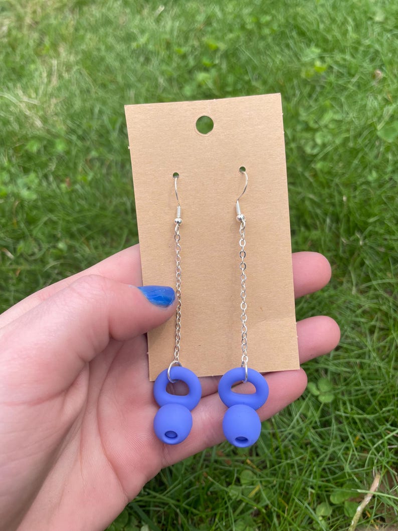 May include: A pair of drop earrings featuring silver-toned metal chains attached to matte blue kettlebell-shaped charms. The earrings are displayed on a rectangular brown cardboard backing card with a circular hole at the top for hanging.