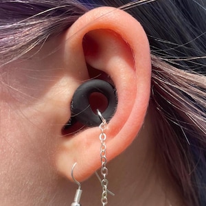 May include: A black circular ear gauge plug inserted into the earlobe, featuring a silver-toned metal chain that connects the plug to a silver-toned fishhook earring wire. The chain hangs in a loop, creating a decorative link between the two pieces.