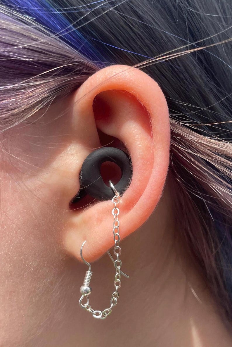 May include: A black circular ear gauge plug inserted into the earlobe, featuring a silver-toned metal chain that connects the plug to a silver-toned fishhook earring wire. The chain hangs in a loop, creating a decorative link between the two pieces.