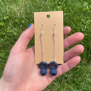 May include: A pair of dangle earrings featuring dark navy blue kettlebell charms suspended from silver-toned metal chains and standard earring hooks, displayed on a rectangular brown cardboard jewelry backing card with a hole at the top.