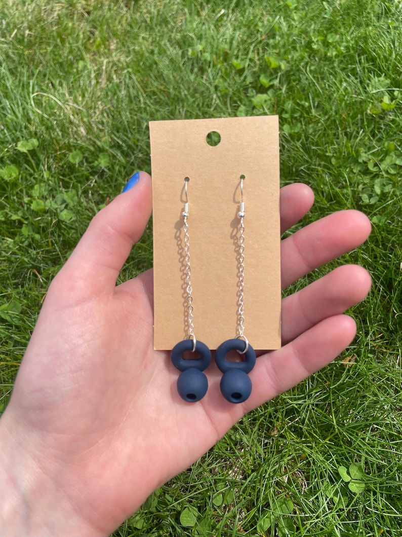 May include: A pair of dangle earrings featuring dark navy blue kettlebell charms suspended from silver-toned metal chains and standard earring hooks, displayed on a rectangular brown cardboard jewelry backing card with a hole at the top.