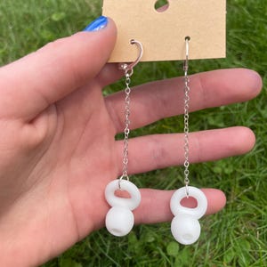 May include: A pair of dangle earrings featuring silver-toned metal chains attached to white, spherical, kettlebell-shaped charms. The earrings are suspended from a rectangular brown cardboard display card with a circular hole at the top center.