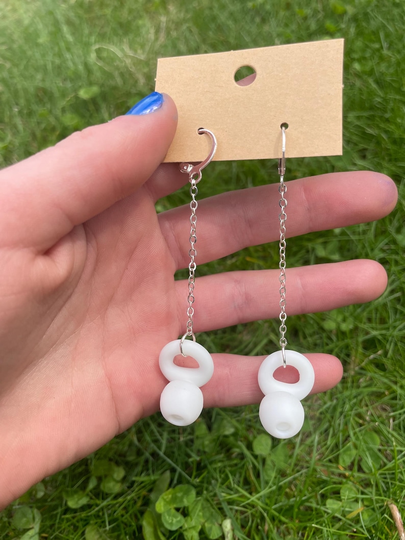 May include: A pair of dangle earrings featuring silver-toned metal chains attached to white, spherical, kettlebell-shaped charms. The earrings are suspended from a rectangular brown cardboard display card with a circular hole at the top center.