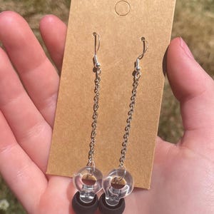 May include: A pair of dangle earrings featuring silver-toned metal chains attached to clear, circular plastic rings. Each earring terminates in a solid matte black spherical bead suspended from the bottom of the clear ring, presented on a brown card.