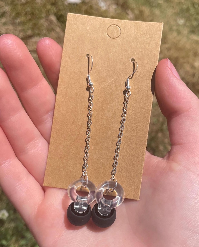 May include: A pair of dangle earrings featuring silver-toned metal chains attached to clear, circular plastic rings. Each earring terminates in a solid matte black spherical bead suspended from the bottom of the clear ring, presented on a brown card.
