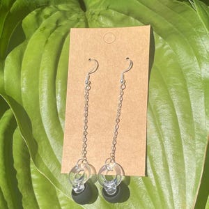 May include: A pair of dangle earrings featuring silver-toned metal hooks and thin chains. Each earring ends with a clear, circular acrylic ring attached to a small, solid black sphere. The earrings are displayed on a rectangular brown card.