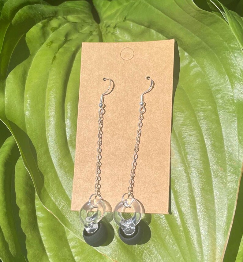 May include: A pair of dangle earrings featuring silver-toned metal hooks and thin chains. Each earring ends with a clear, circular acrylic ring attached to a small, solid black sphere. The earrings are displayed on a rectangular brown card.