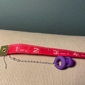 May include: A purple earplug earring featuring a silver-toned metal chain and a standard hook finding. The earring is positioned alongside a red measuring tape, which indicates the total length of the piece is approximately 3.5 inches long.